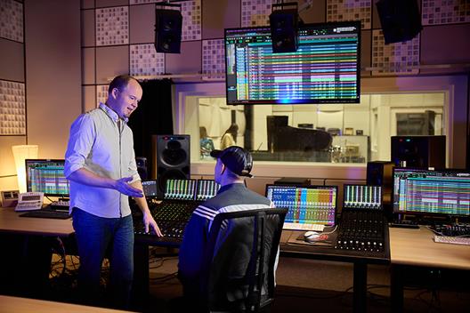 Insight into the recording studio at Düsseldorf University of Applied Sciences. The room is equipped with a large mixing desk and lots of technology. In the background you can see a large window with a view of the recording room. A young man is sitting at the mixing desk talking to another young man standing in front of the desk.